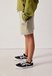 Persoon die beige korte broek met tekst 'American Vintage', een groene trui en zwart-witte sneakers draagt, staand in zijaanzicht.