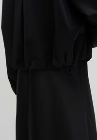 Close-up of a black jacket with elastic hem and visible drawstring, layered over a black garment, plain light gray background.