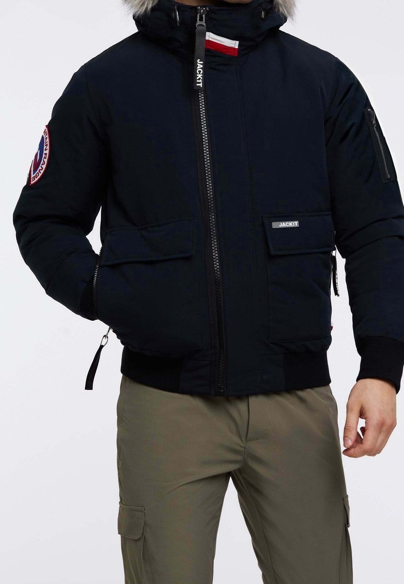 Veste bleu marine foncé avec capuche bordée de fourrure, deux poches avant, fermeture éclair et détails de logo. Le matériau semble doux et durable.