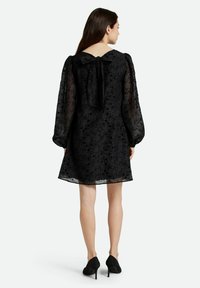 Vestido negro con mangas largas y transparentes, patrón floral y un lazo en la parte posterior. El dobladillo está ligeramente por encima de la rodilla, complementado con tacones negros.