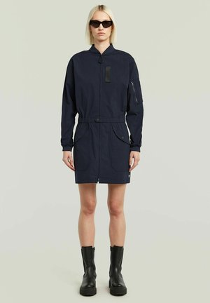 Femme portant une robe longue à manches longues bleu marine avec fermeture éclair et poches, des bottes noires mi-mollet et des lunettes de soleil noires, debout devant un fond uni.