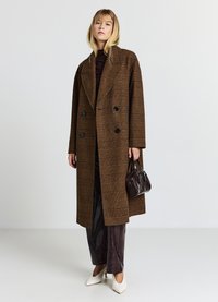 Brauner kariierter doppelt-knöpfbarer Übermantel mit großen Revers, schwarzen Knöpfen, kombiniert mit dunkelbraunen Hosen und einer kleinen schwarzen Handtasche. Weiße Absätze.