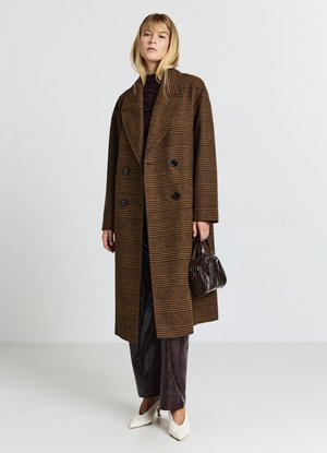 Manteau croisé à carreaux marron, grands revers, boutons noirs, associé à un pantalon marron foncé et un petit sac à main noir. Escarpins blancs.