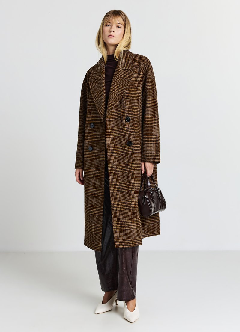 Brauner kariierter doppelt-knöpfbarer Übermantel mit großen Revers, schwarzen Knöpfen, kombiniert mit dunkelbraunen Hosen und einer kleinen schwarzen Handtasche. Weiße Absätze.