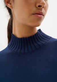 Marinblå stickad tröja med hög ribbad halsringning, tillverkad av slät tyg. Nära bild som framhäver kragen och sömnadsdetaljer.