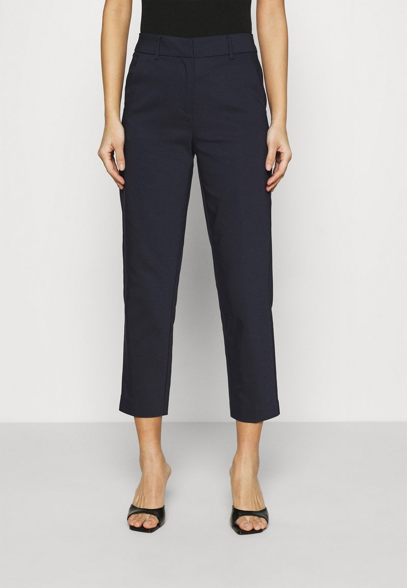 Femme portant un pantalon bleu marine court à jambe droite et des sandales à talons ouvertes noires, debout devant un fond uni.