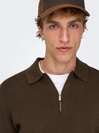 Polo marrone con zip, collo a risvolto, tessuto strutturato e dettaglio con cerniera argentata; indossata sopra una t-shirt bianca e abbinata a un cappello marrone.