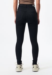 Svarta högmidjade skinny jeans med en slät yta, bakfickor och en märkesetikett vid midjan, tillsammans med ljusa sneakers.