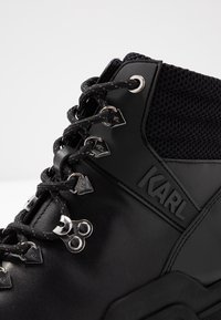 Bota negra de tobillo con cuero y tela texturizada, con herrajes plateados, ganchos triangulares para los cordones y un logotipo destacado en el lateral.