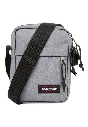 Pequeño bolso cruzado gris con correa negra ajustable, bolsillo frontal con cremallera y parche con logo de Eastpak en la parte delantera.