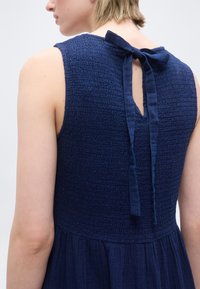 Άτομο που φοράει αμάνικο ναυτικό μπλε φόρεμα με υφή, με δεμένη φιόγκο στο πίσω μέρος του λαιμού και λεπτομέρεια με φούστα που μαζεύει.