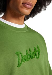 Felpa verde con un logo ricamato verde a trama. Collo a crew, tessuto morbido e vestibilità rilassata. Nessun motivo visibile o dettagli aggiuntivi.