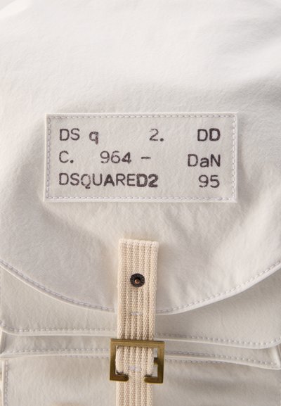 Άσπρη υφασμάτινη ετικέτα με μαύρο κείμενο "DSQUARED2" ραμμένη στην κρεμ φλάπα τσάντας ασφαλισμένη με μπεζ λουρί και ορειχάλκινη αγκράφα.