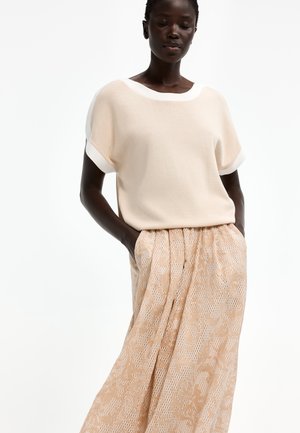 Frau trägt ein cremefarbenes Kurzarmshirt und einen hoch taillierten beigen Spitzenrock mit Händen in den Taschen vor weißem Hintergrund.