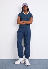 Marina atletiska byxor med elastiska ärmslut, i kombination med en turkos t-shirt med vit krage och broderad logga, kompletterad med en lavendelfärgad keps.