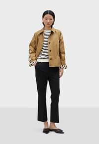 Kaki-Jacke mit Knöpfen, gestreifter Pullover, schwarze Hose und schwarze spitze Ballerinas. Pünktchenakzente an den Ärmeln des Pullovers.