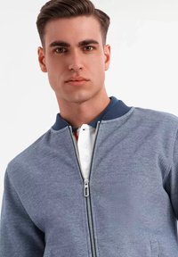 Chaqueta bomber con cremallera en tejido azul texturizado, con un collar navy rígido, superpuesta sobre una camiseta polo blanca con detalles de botones.