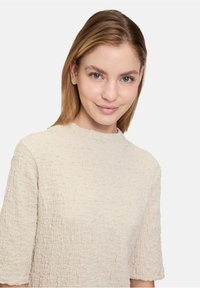 Haut texturé beige avec un col montant et des manches courtes, présentant un motif de tissu froncé. Surface de tissu lisse visible.