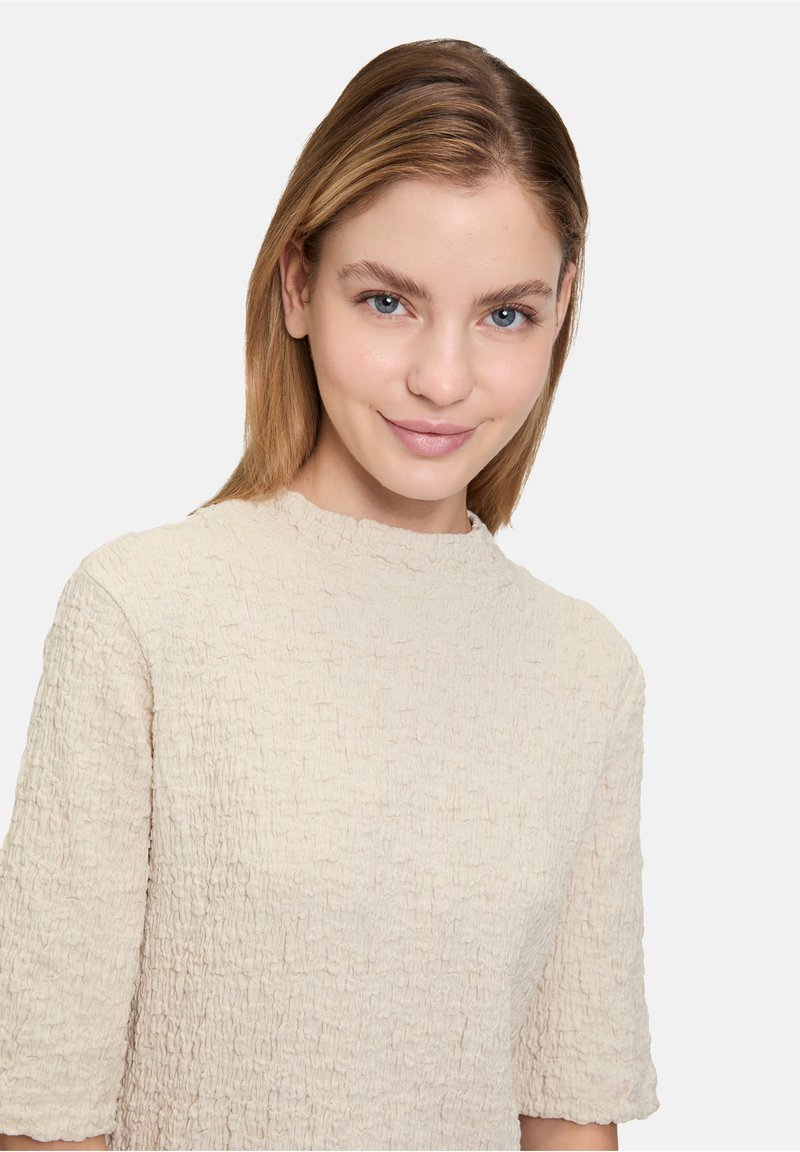 Haut texturé beige avec un col montant et des manches courtes, présentant un motif de tissu froncé. Surface de tissu lisse visible.