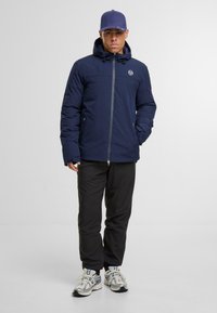 Marineblauwe gewatteerde jas met ritssluiting, verstelbare capuchon en logo-opdruk op de borst; gecombineerd met zwarte broeken en grijze sneakers met blauwe accenten.