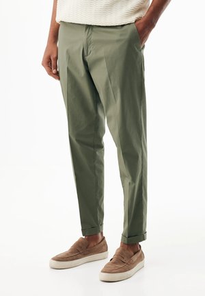 Osoba w oliwkowozielonych spodniach z mankietem, z rękami w kieszeniach, ubrana w brązowe zamszowe buty wsuwane z białymi podeszwami.