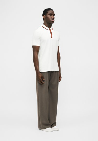 Άνδρας που φοράει λευκό πόλο με κοντά μανίκια και πορτοκαλί και μαύρη διακόσμηση, συνδυασμένο με φαρδιά καφέ παντελόνια και λευκά αθλητικά παπούτσια, στέκεται ακίνητος.