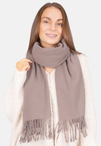 Jeune femme souriante, portant un pull en tricot épais beige et une écharpe taupe épaisse avec des bords frangés, tenant un côté de l'écharpe.