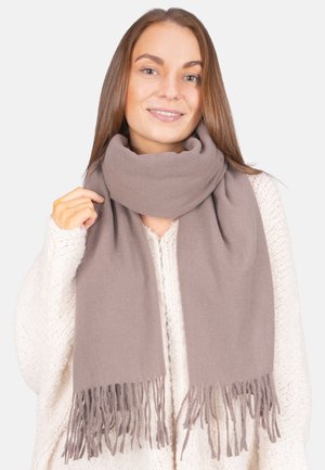 Jeune femme souriante, portant un pull en tricot épais beige et une écharpe taupe épaisse avec des bords frangés, tenant un côté de l'écharpe.