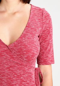 Haut cache-cœur rouge côtelé à manches courtes, avec un décolleté en V profond et un détail de nouage sur le côté. Le tissu présente un motif texturé et moucheté.