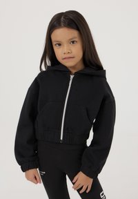 Felpa corta nera con zip frontale, grandi tasche laterali e vita elasticizzata. Abbinata a leggings neri con logo bianco.