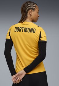 Maglia sportiva gialla con maniche lunghe nere. Testo "DORTMUND" in grassetto lettere nere sul retro. Tessuto liscio, design aderente.