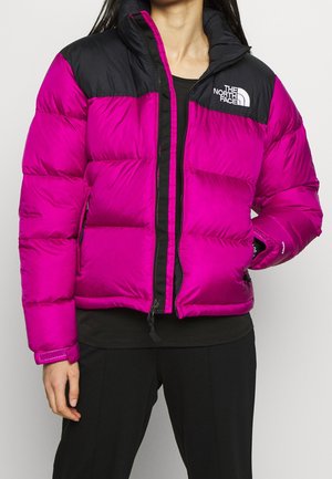 Personne portant une doudoune North Face rose vif et noire sur un pantalon noir, avec une main dans la poche de la veste.