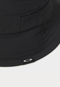 Fekete, könnyű anyagból készült sapka, perforált dizájnnal, varrt részletekkel és egy kis márkázott címkével, amelyen fehér logó található.