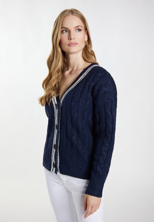 Vrouw met lang blond haar die een marineblauwe cable knit cardigan draagt met witte biezen en zwarte knopen, gecombineerd met een witte broek, kijkt opzij.