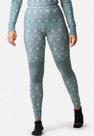 Osoba nosící termolegíny s tyrkysovým vzorem a odpovídající dlouhý rukáv s černými ponožkami, stojící před bílým pozadím.