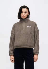 Brązowy polarowy sweter z wysokim kołnierzem, suwakiem do �ćwiartki oraz logo „GAP”. Posiada raglanowe rękawy i luźny krój. Miękka faktura.