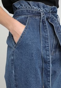 Mano descansando en el bolsillo de unos pantalones de mezclilla azul de talle alto con un cinturón atado y una parte superior negra visible a la altura de la cintura.