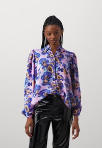 Blusa de manga larga en tejido púrpura con un patrón floral en azul y amarillo. Cuenta con un cuello tipo polo y mangas ligeramente abullonadas.