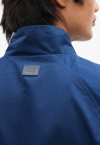 Ναυτικό μπλε μπουφάν με καλοσχηματισμένο γιακά, παρουσιάζοντας μια δερμάτινη επένδυση με λογ ότυπο πίσω. Ο ιστός φαίνεται λείος με ματ φινίρισμα.