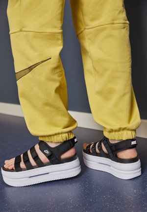 Czarne sandały na platformie z wieloma paskami, z białą podeszwą i brandingiem Nike. W połączeniu z żółtymi joggerami, prezentującymi luźny krój.