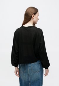 Kvinne med langt brunt hår som har på seg en løs svart genser med lange ermer og et blått denimskjørt, sett bakfra mot en ensfarget bakgrunn.