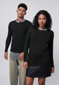 Zwarte truien met ribbels aan de mouwen en een "ICONIC" logo op de kraag. Model draagt het met vervaagde denim en een zwarte leren rok.
