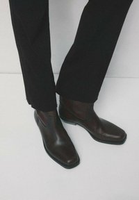 Osoba w czarnych spodniach o prostych nogawkach i ciemnobrązowych skórzanych botkach Chelsea na jasnoszarej podłodze.