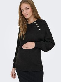 Nėščia moteris dėvi juodą megztinį su sidabrinėmis sagomis ant peties, švelniai laikydama pilvą, stovėdama priešais šviesų, paprastą foną.