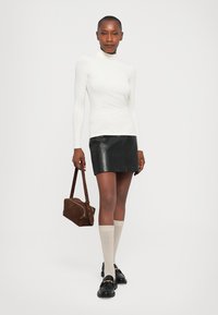 Bijela duga majica s turtleneck-izrezom uparena s crnom mini suknjom od kože i bež čarapama do koljena. Manekenka drži smeškastu torbu.