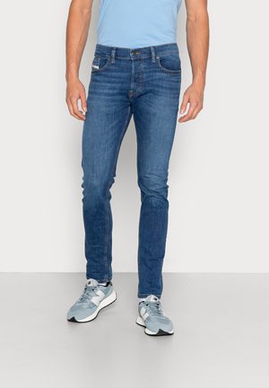 Homme portant un jean slim bleu et des baskets New Balance bleu clair, debout devant un fond blanc uni.