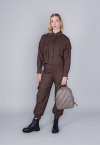 Camisa corta marrón con dos bolsillos frontales y pantalones ajustados a juego, combinados con botas negras con cordones y una mochila acolchada gris claro.