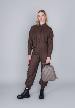 Bruine cropped shirt met twee voorke pockets en bijpassende tapered broek, gecombineerd met zwarte veterschoenen en een lichtgrijze gewatteerde rugzak.
