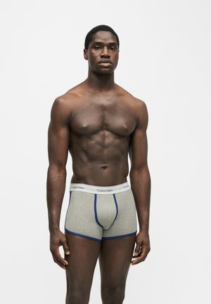 Atletický muž stojaci na jednofarebnom pozadí, oblečený v sivých boxerkách s modrým lemom a bielou páskou s logom Calvin Klein.