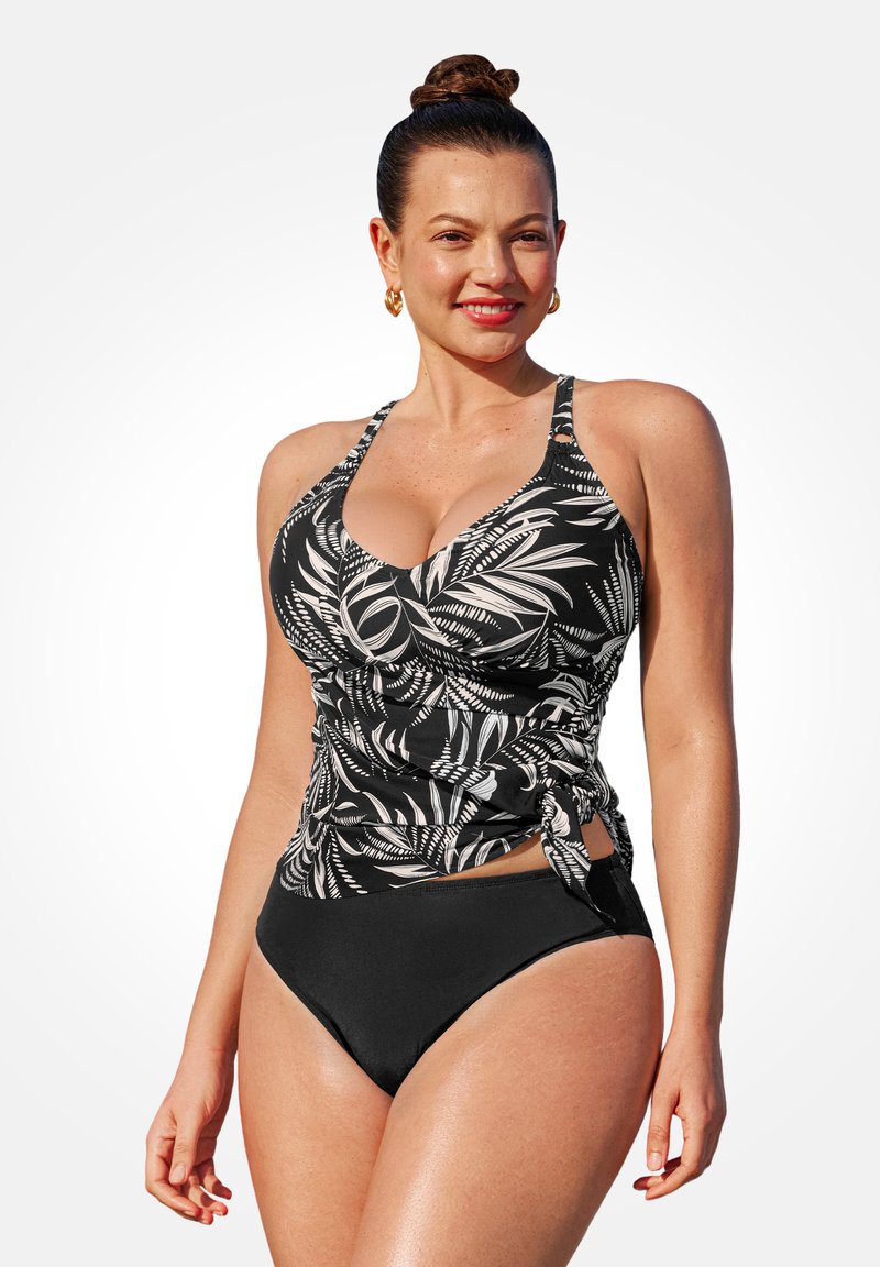 Donna sorridente con capelli raccolti in un bun, che indossa un top tankini con stampa foglia nera e bianca con fiocco laterale e costumi da bagno neri semplici.